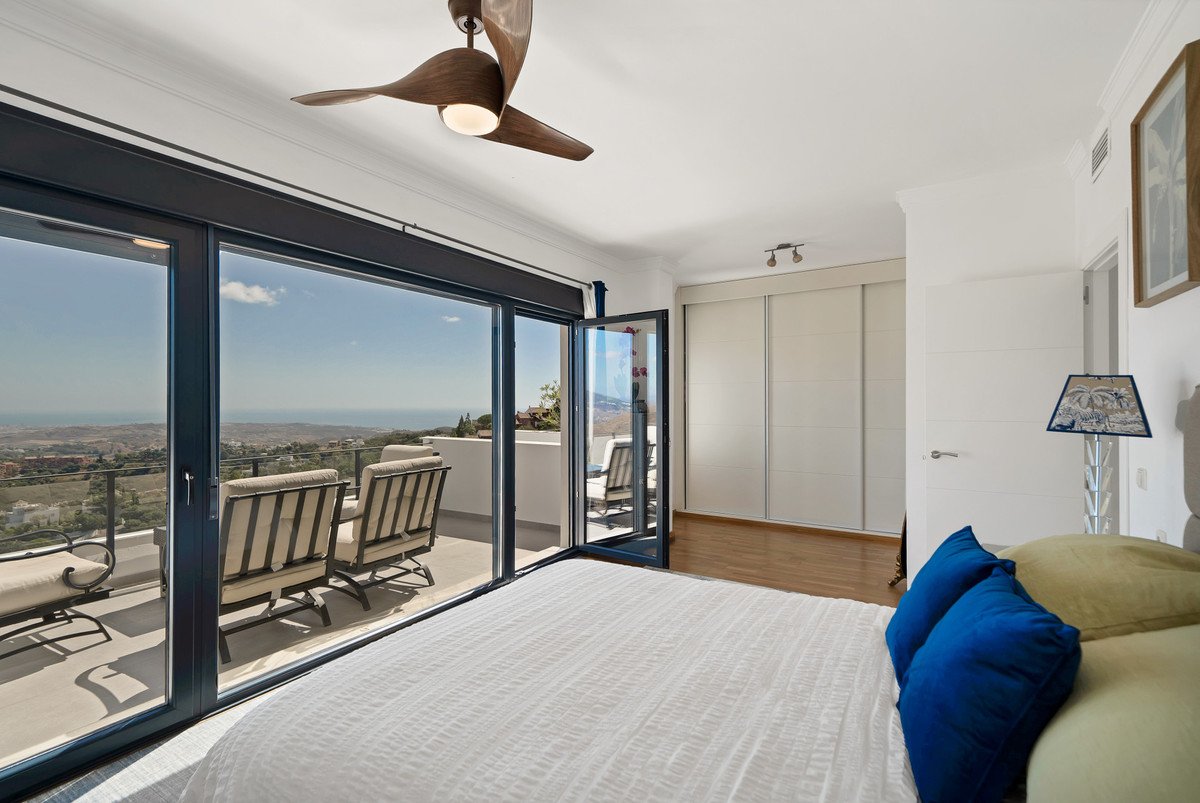 Bedroom with large windows opening to a balcony with seating and a scenic sea and landscape view.