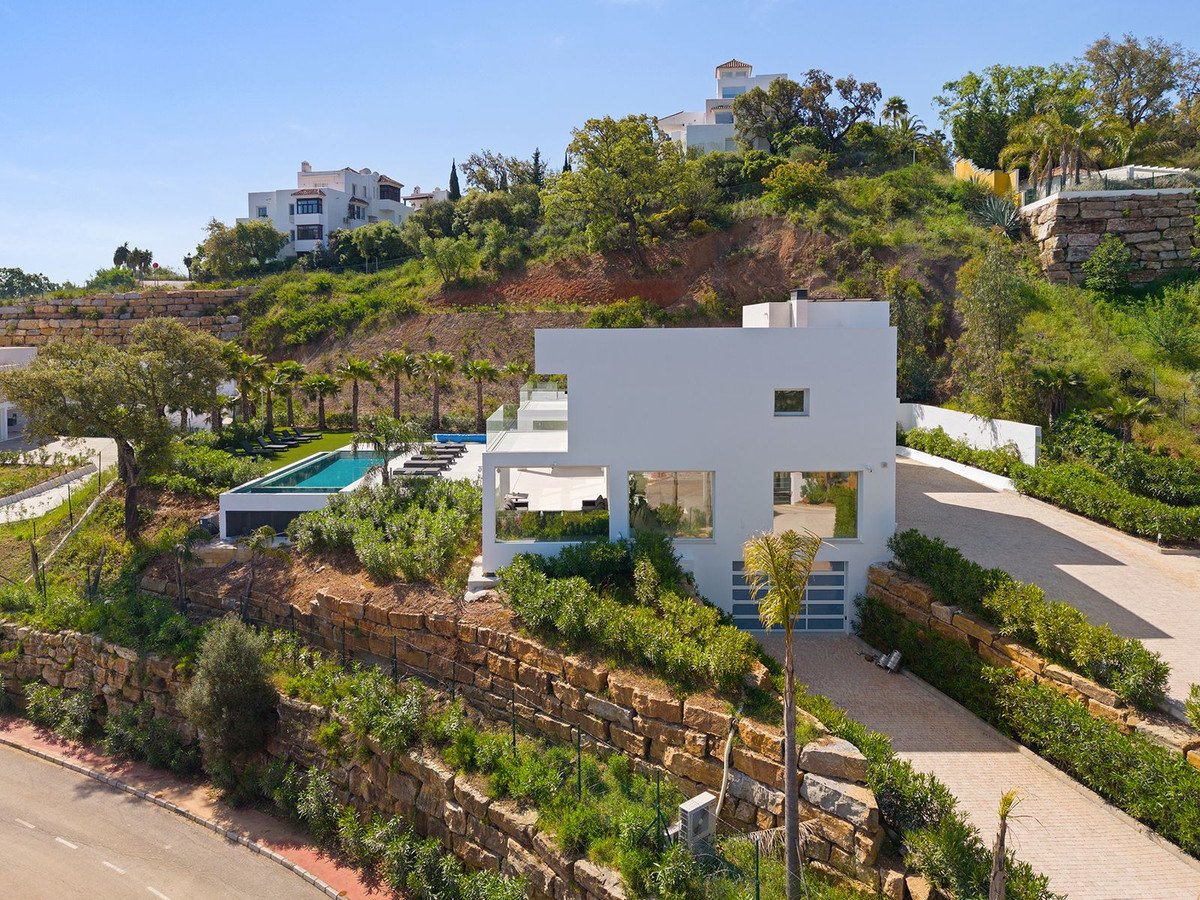 Villa blanca moderna con piscina y tumbonas, en colina con vegetación exuberante y mar lejano.