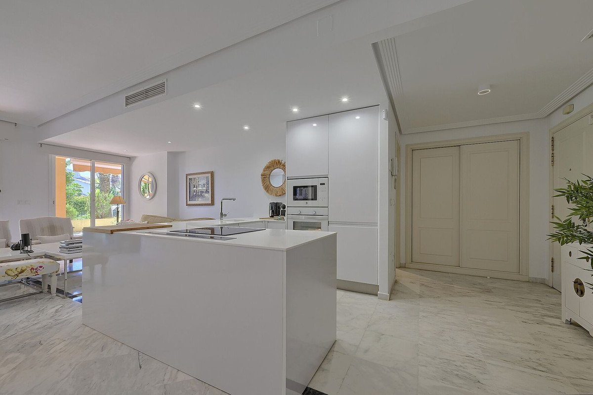 Modern white kitchen with island, integrated appliances, and views to the living area and outdoors through large windows.