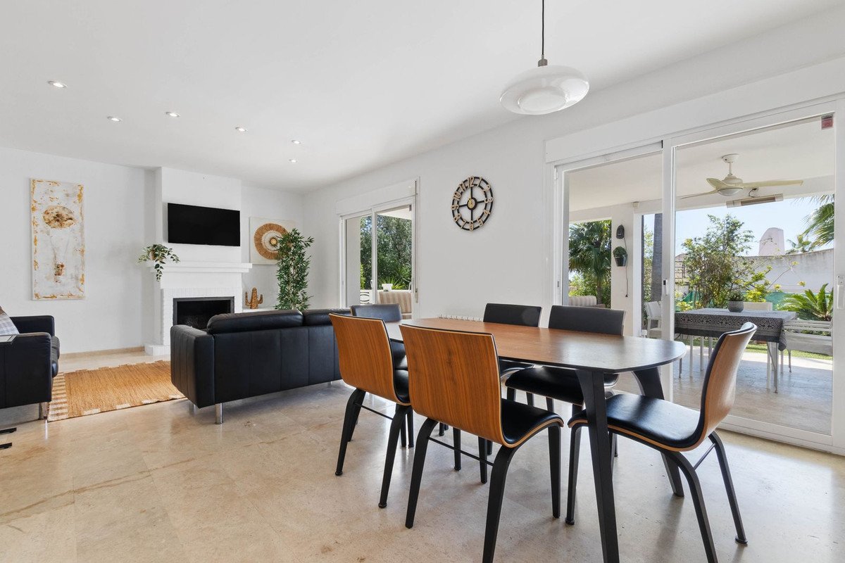 Modern living and dining area with black sofas, wooden chairs, fireplace, and sliding doors opening to a sunny patio with outdoor seating.