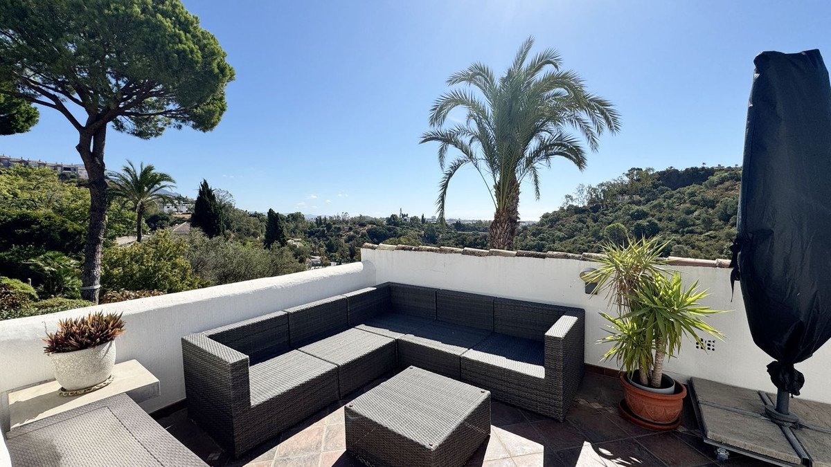 Rattan lounge seating on a terrace overlooking lush green hills and palm trees under a clear blue sky.
