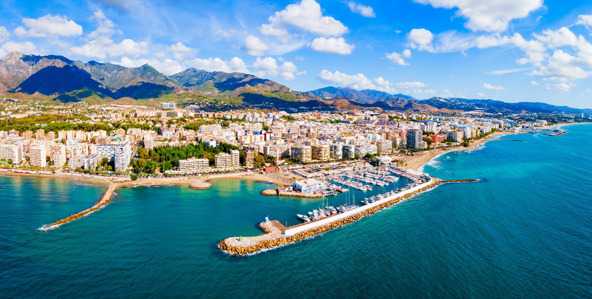 Aerial view of a coastal city with a marina, mountains, and clear blue water.