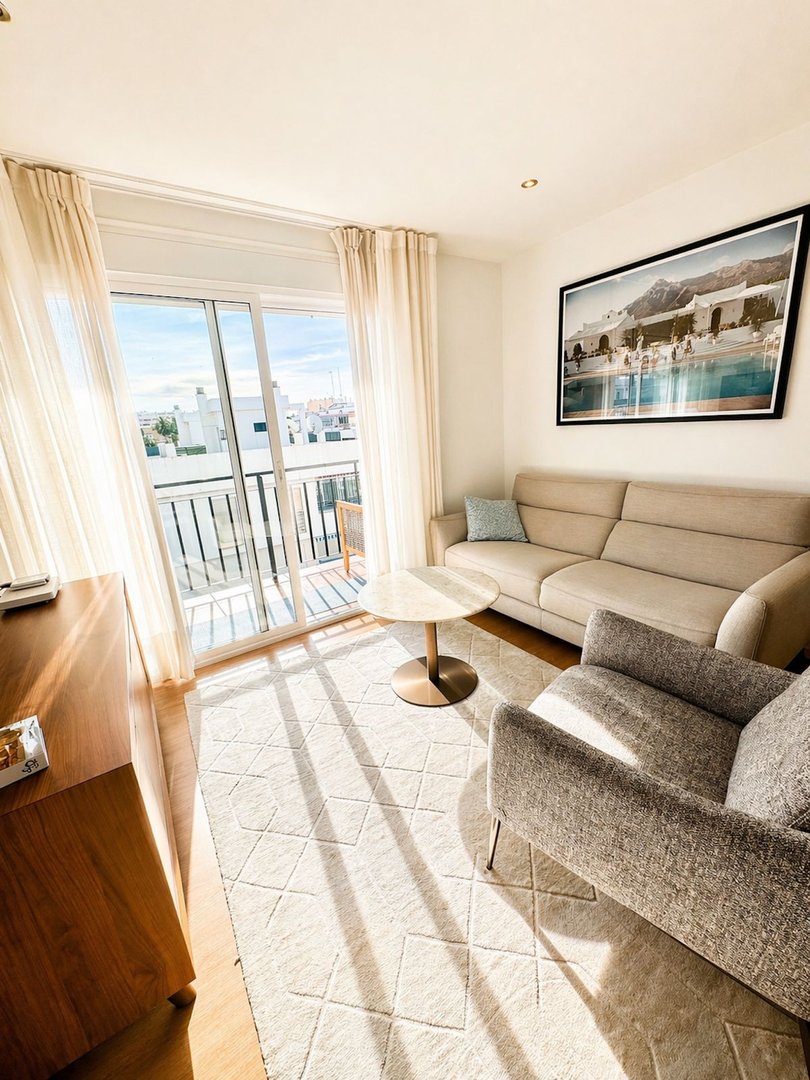 Modern living room with beige sofa, armchair, coffee table, and balcony with city view.