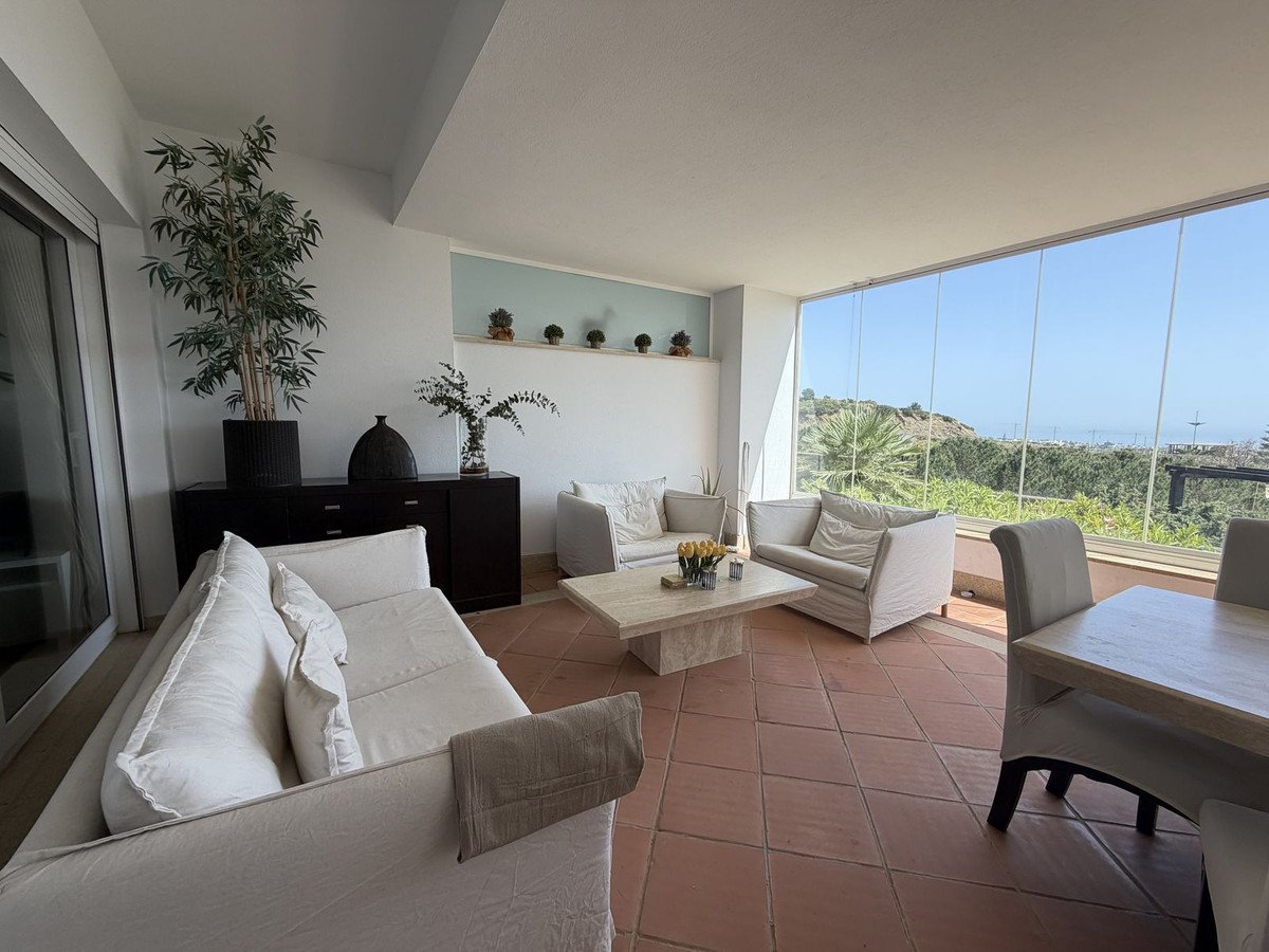 Modern living room with sofas, coffee table, and dining set, featuring a glass-enclosed balcony with stunning sea and landscape views.