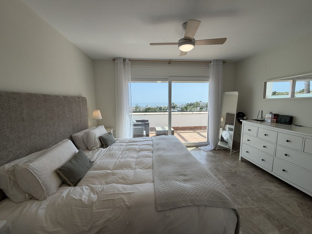 A spacious bedroom with a large bed, leading to a terrace with a sea view, outdoor seating, and lush greenery.