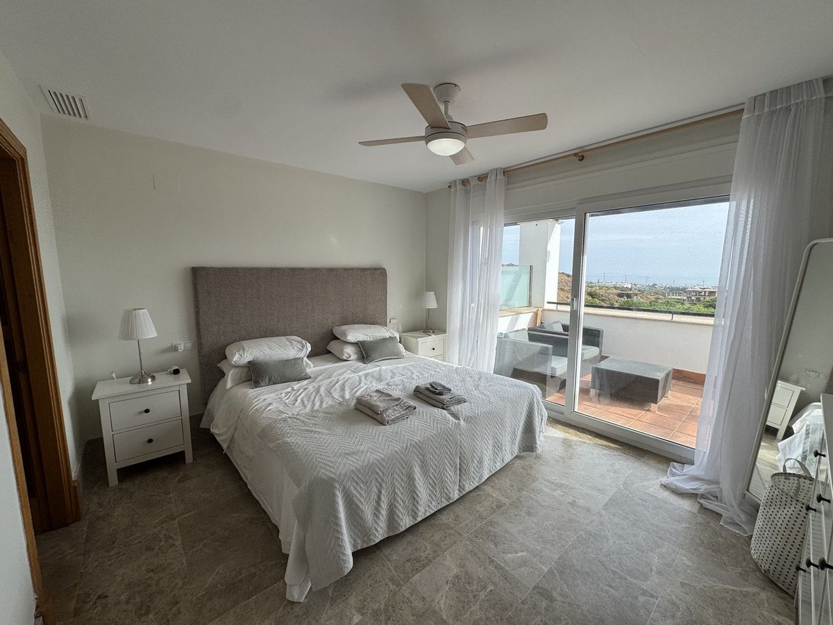 A spacious bedroom with a large bed, nightstands, and a ceiling fan, featuring a balcony with outdoor seating and a distant sea view.