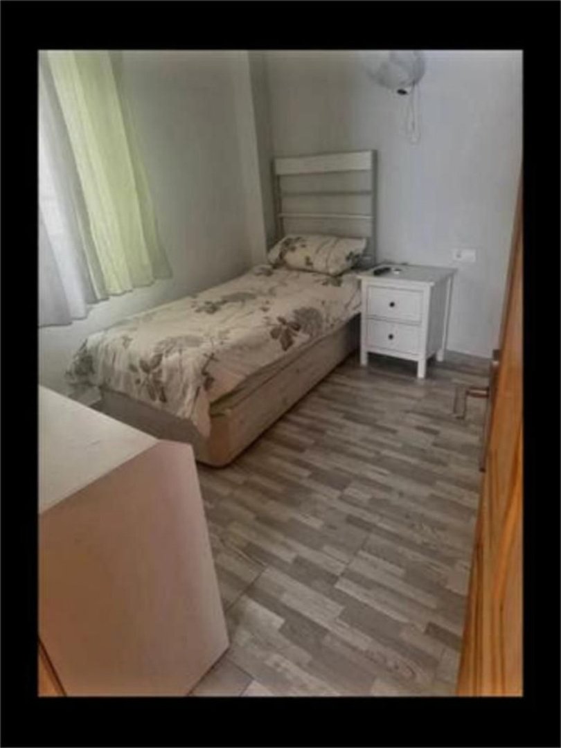 A small bedroom featuring a single bed with floral bedding, a white nightstand, and light grey wood-look flooring. Light streams in from a window with sheer.