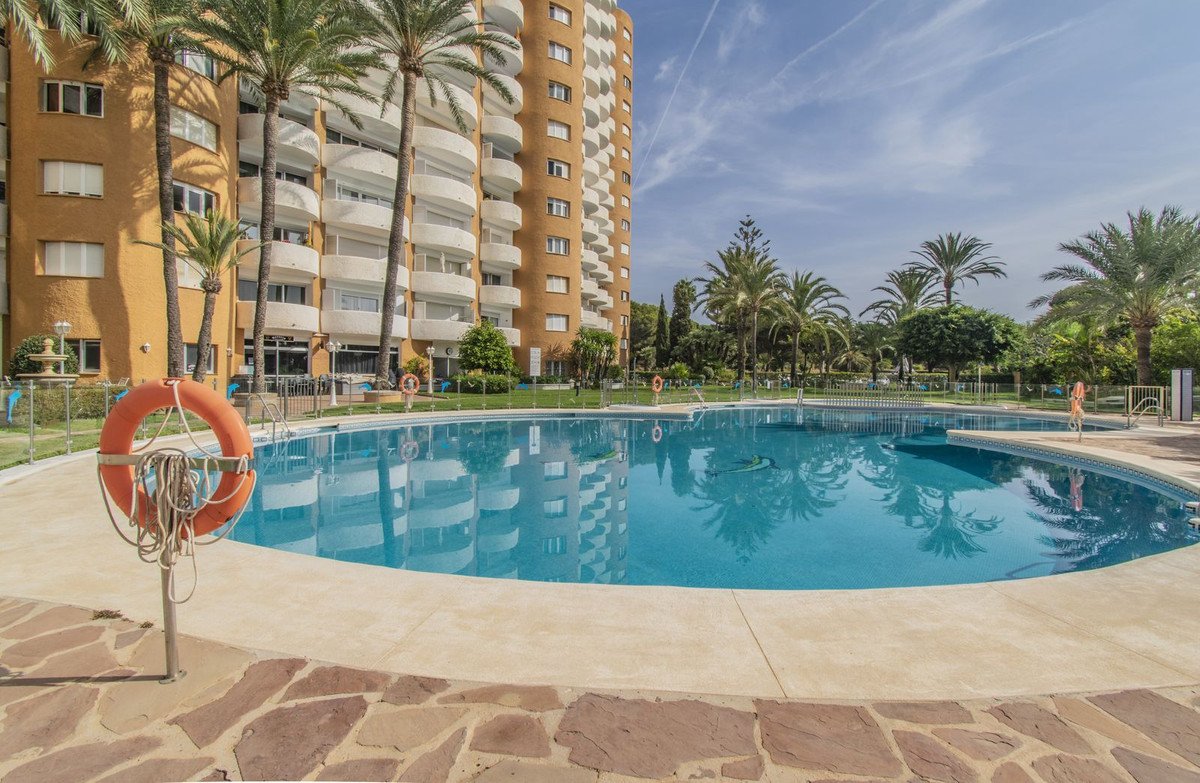 View of a large swimming pool and apartment building with palm trees in a sunny garden setting.