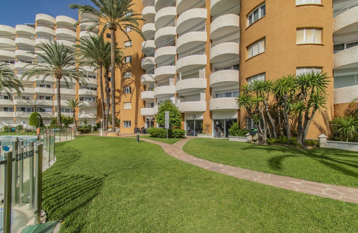Apartment building with balconies, palm trees, green lawn, pathway, and swimming pool area.