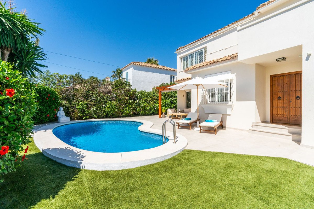 A private swimming pool and terrace with lounge chairs, umbrellas, and lush green lawn, in front of a white villa under a clear blue sky.