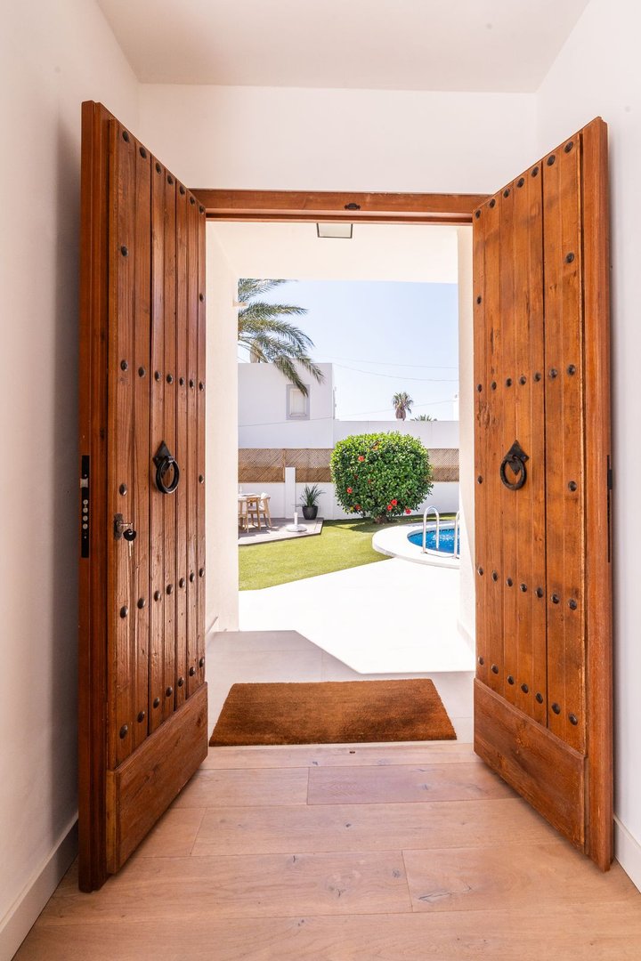 View through open wooden doors to a sunny garden with a swimming pool, lawn, and outdoor seating area.