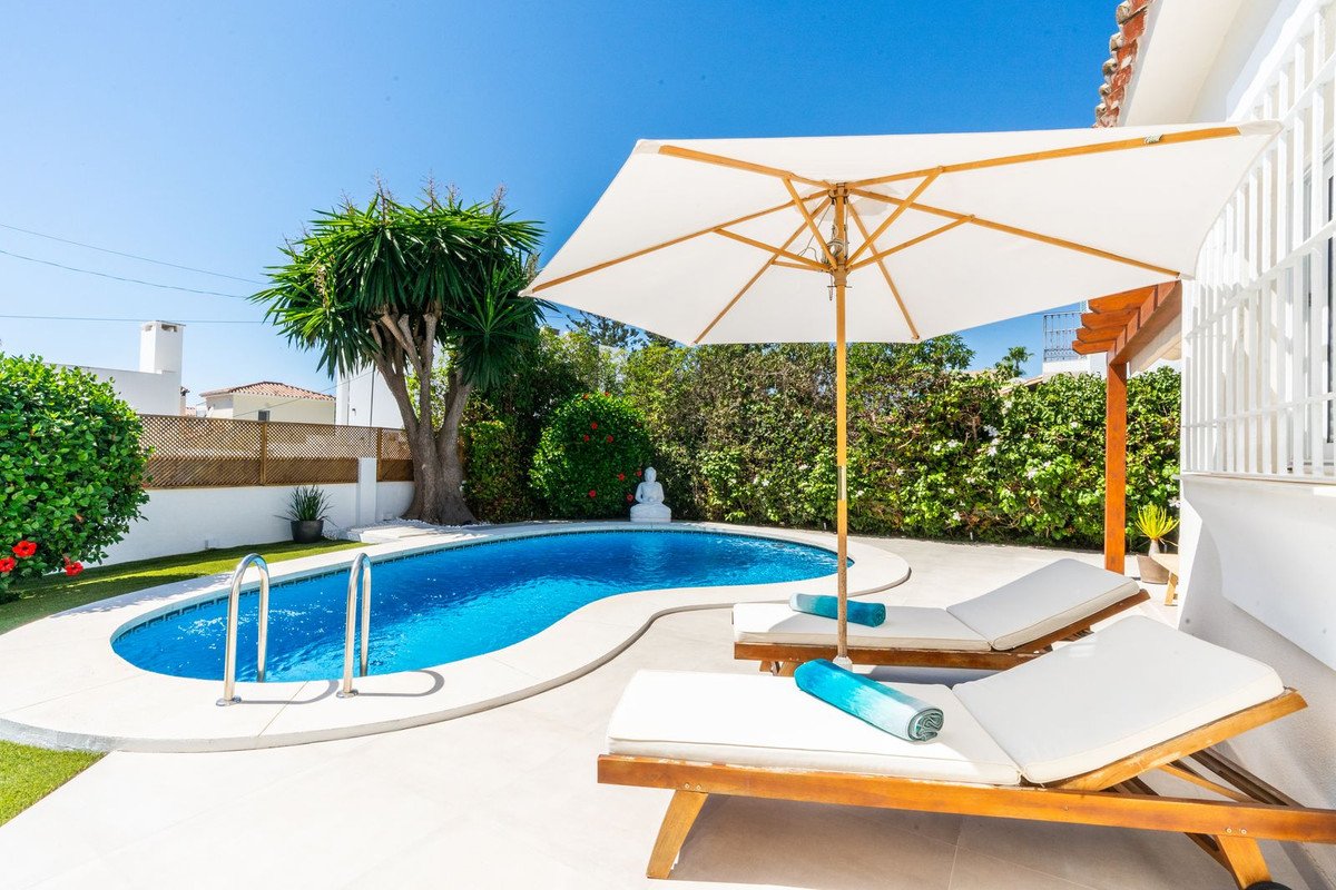 A kidney-shaped swimming pool with lounge chairs and a white umbrella on a sunny day, surrounded by lush greenery.