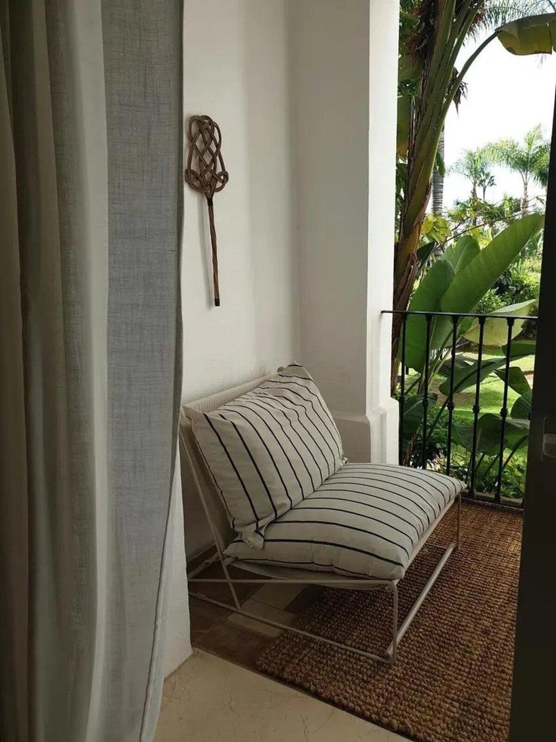 A cozy balcony with a chair and cushions, offering a lush view of tropical plants and palm trees.