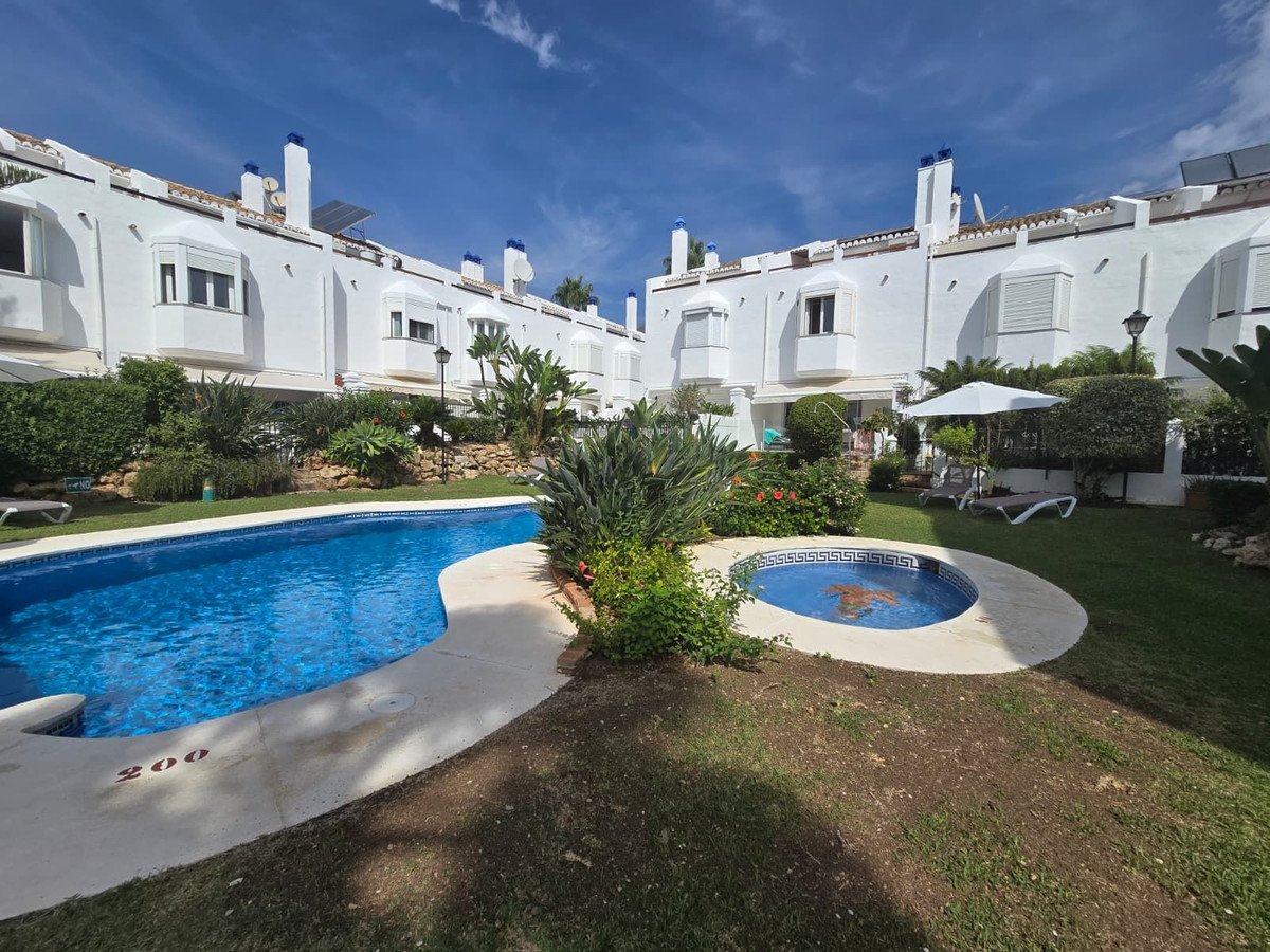 Exterior view of white townhouses with a curved swimming pool, a small jacuzzi, and a lush green garden on a sunny day.