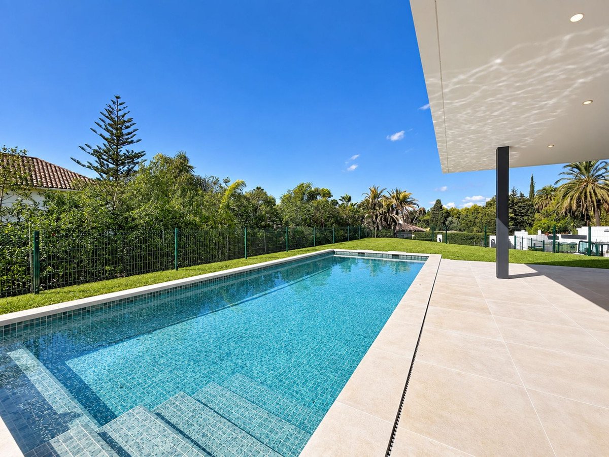 A modern swimming pool with a tiled terrace and a lush green garden on a sunny day.