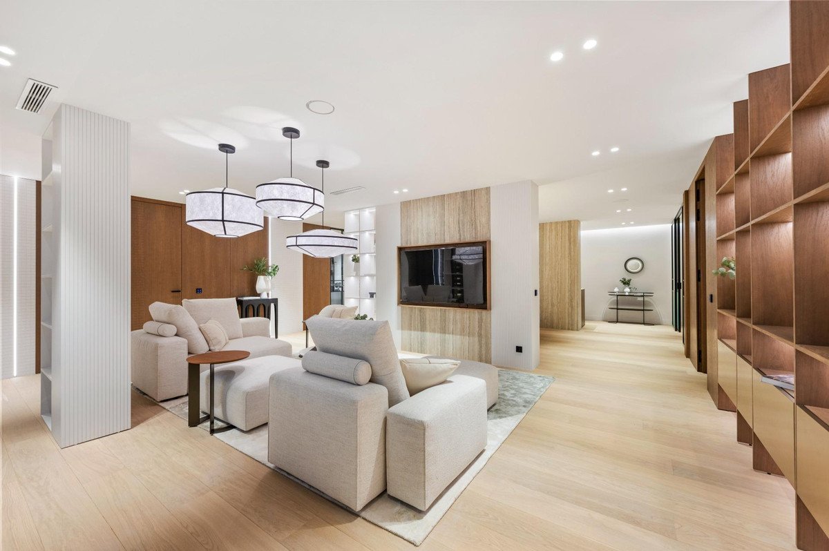 A modern living room with light-colored sofas, wood-paneled walls, decorative lighting, and a wall-mounted television.
