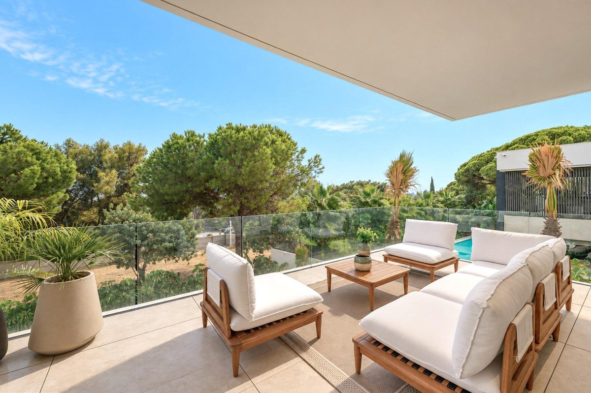 Modern terrace with white sofas and coffee table, overlooking lush green trees and a swimming pool under a blue sky.