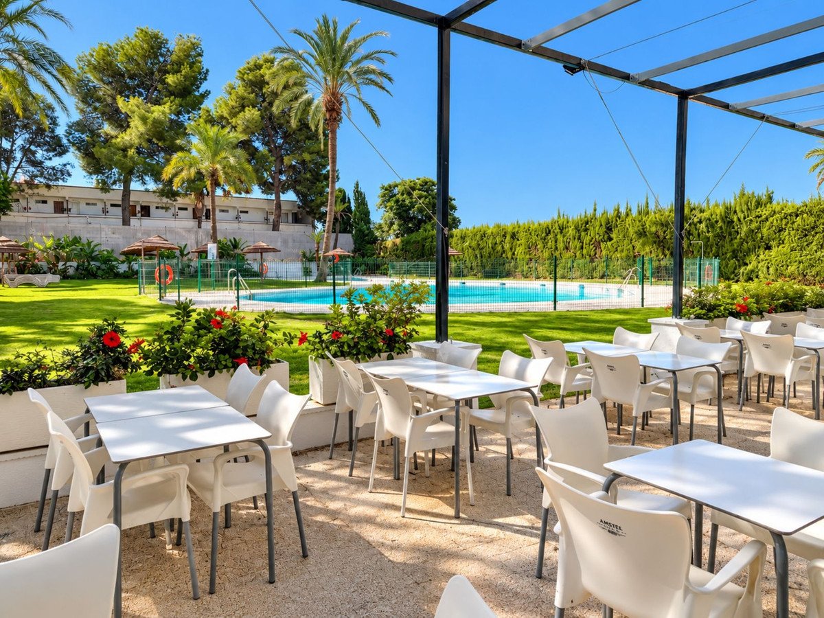 Outdoor dining area with white tables and chairs, overlooking a swimming pool and lush green garden with palm trees.