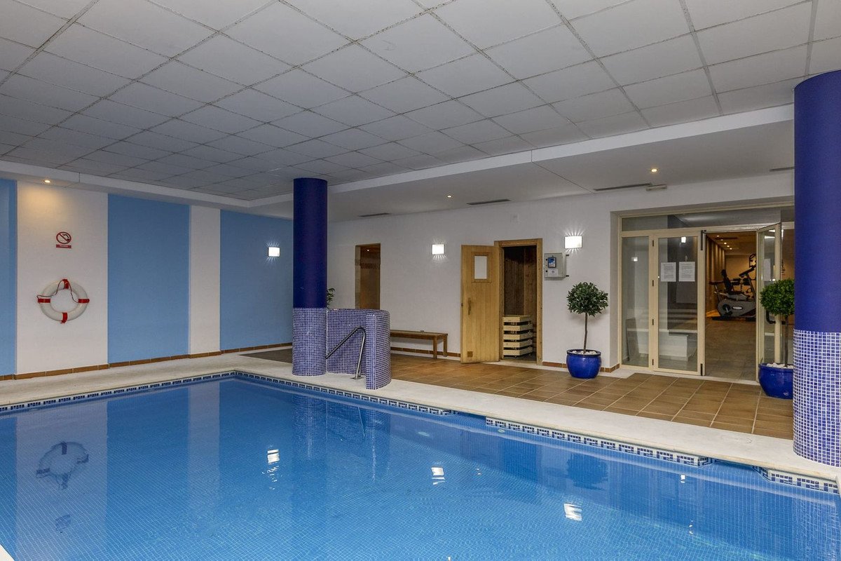 Indoor swimming pool with blue tiles, a wooden sauna, and gym equipment visible through a glass door.