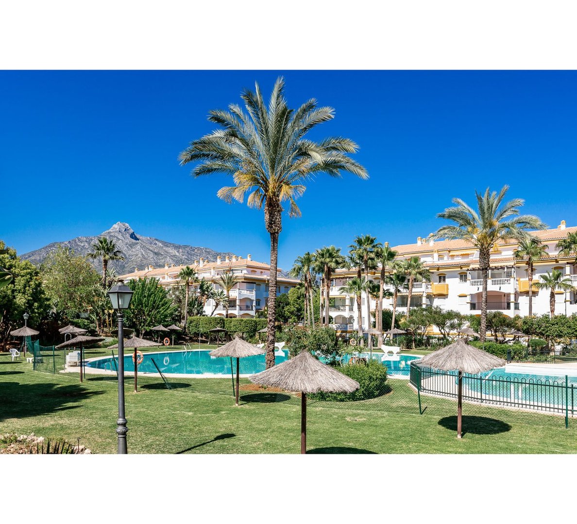 Resort complex with swimming pools, palm trees, sun umbrellas, and a mountain visible in the background under a clear blue sky.