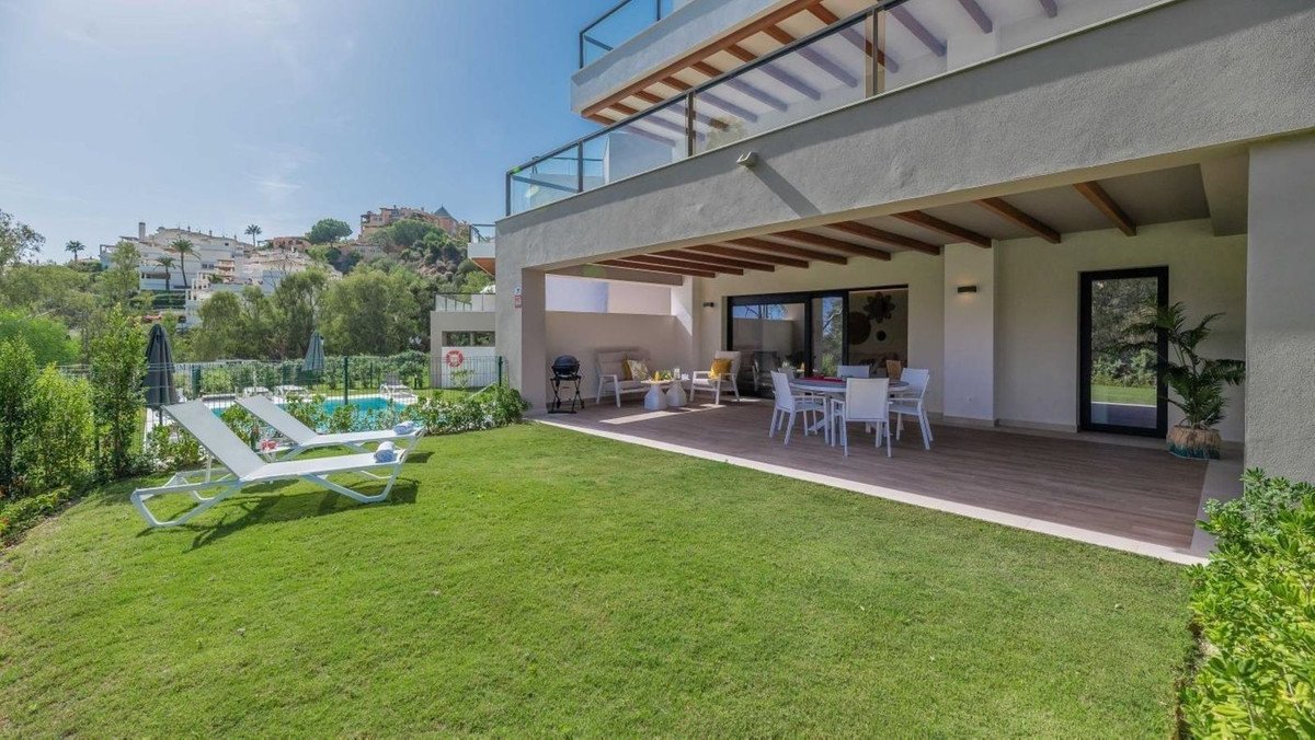 A modern villa terrace with outdoor dining, lounge area, and a green lawn leading to a swimming pool with lounge chairs.