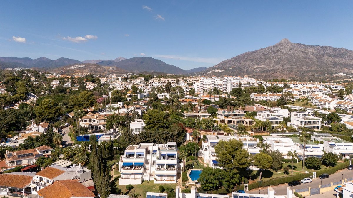 Aerial view of a residential complex with white buildings, greenery, and mountain backdrop.