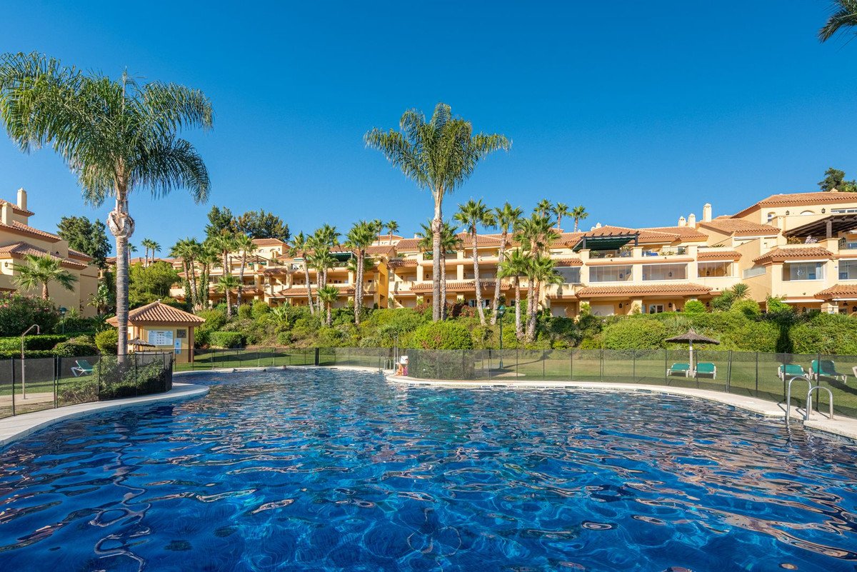 A sparkling blue swimming pool in front of a building complex with palm trees under a clear blue sky.