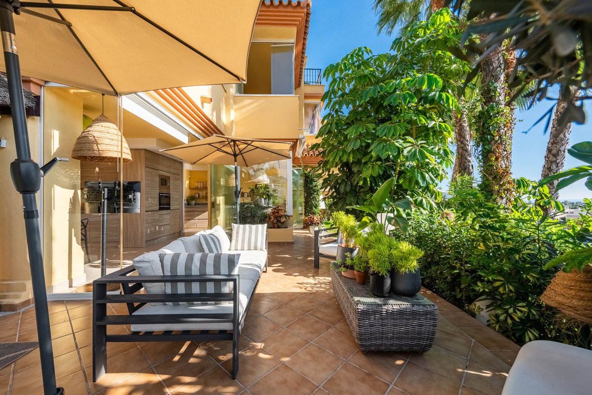Terrace with outdoor kitchen, seating area, umbrellas, and lush green plants on a sunny day.