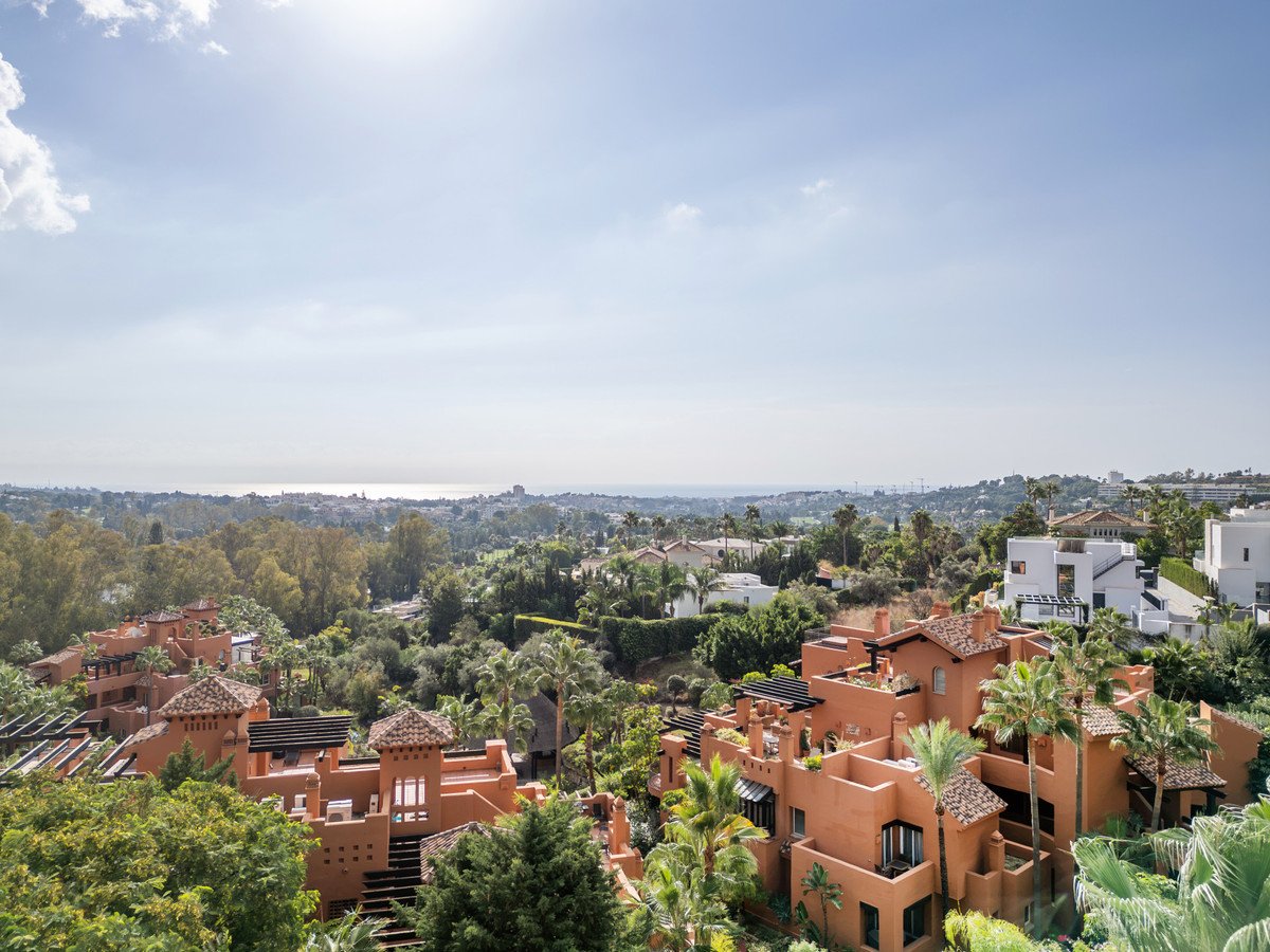 An expansive view of terracotta-roofed houses and lush greenery with a distant sea horizon under a bright sky.