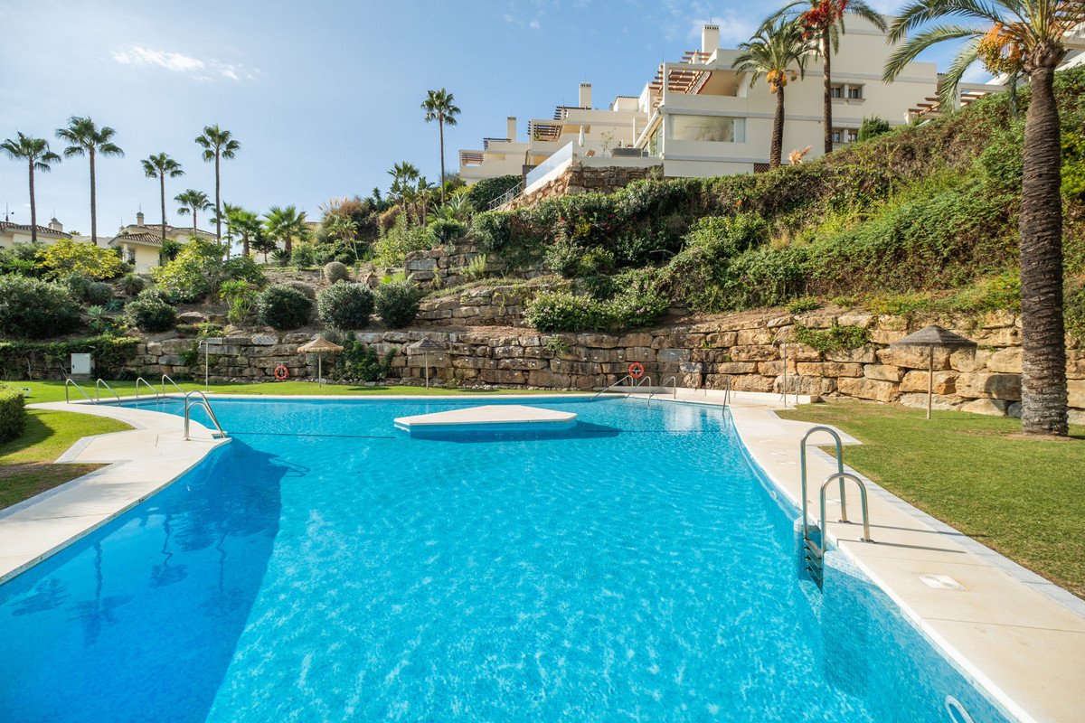 A resort-style community swimming pool with palm trees and modern apartments on a hillside, under a clear blue sky.