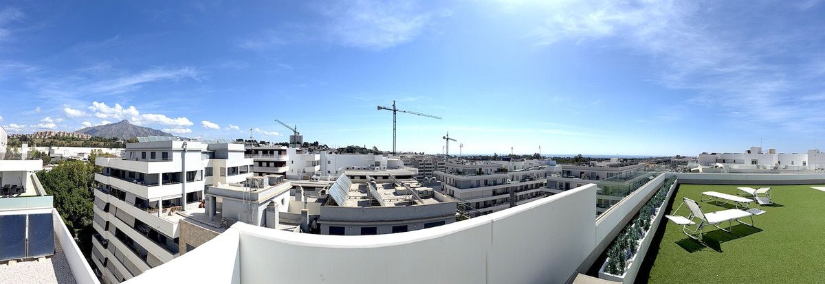 Rooftop terrace with artificial grass, lounge chairs, and a panoramic view of buildings and distant mountains under a blue sky.