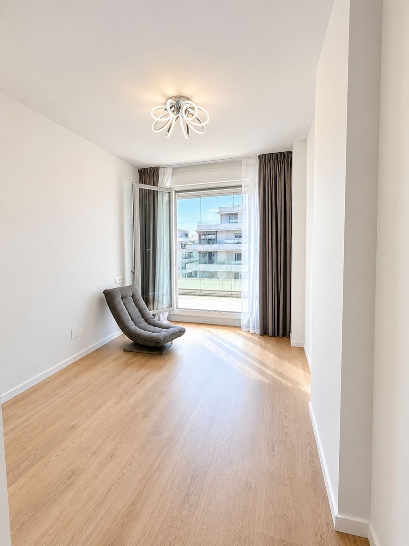 Empty room with a modern grey chair, large window with curtains, and a balcony view of apartment buildings.