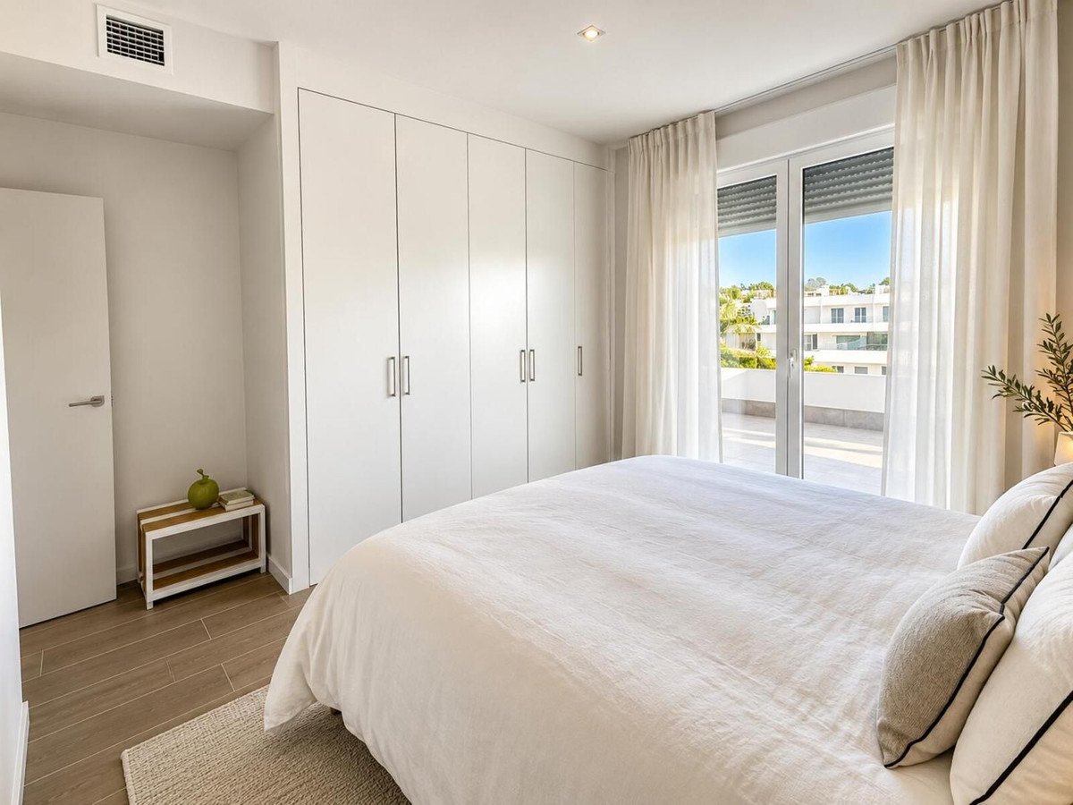 A bright bedroom with a large white bed, built-in wardrobes, and patio doors opening onto a balcony with a view of modern buildings.