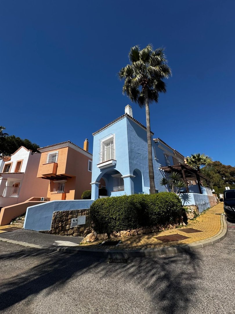 A bright blue house with a balcony and palm tree in front, set against a clear blue sky. Orange house to the left.