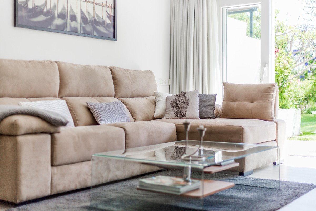 A bright living room with a beige sofa, glass coffee table, and large windows overlooking greenery.