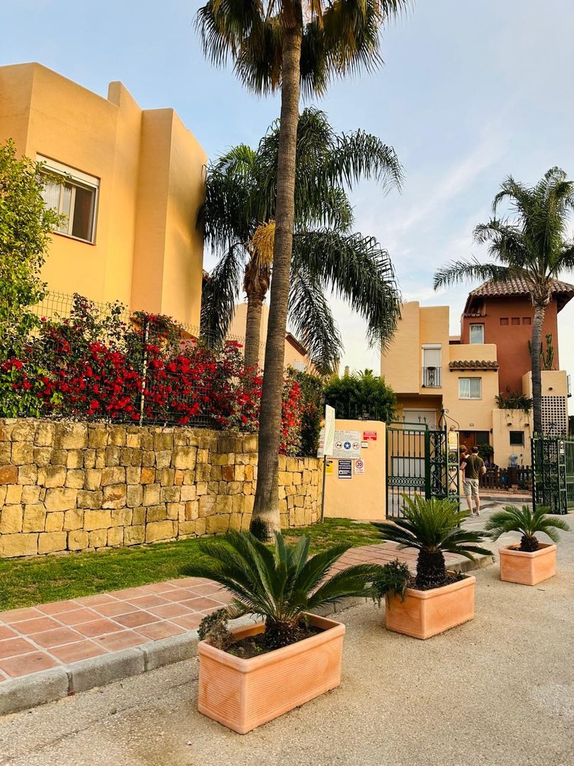Exterior view of a yellow Spanish villa with palm trees and potted plants lining the street.