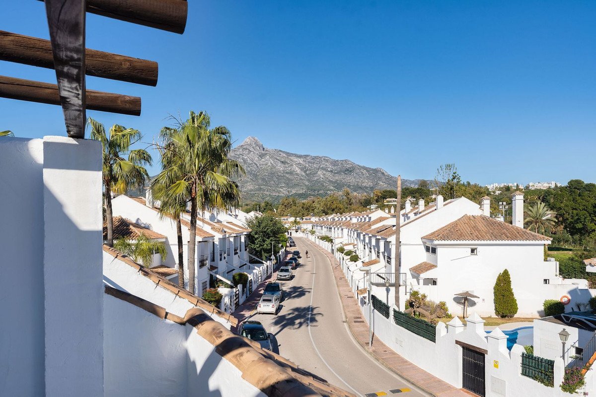 View of a street with white houses, palm trees, and mountains in the distance under a clear blue sky.