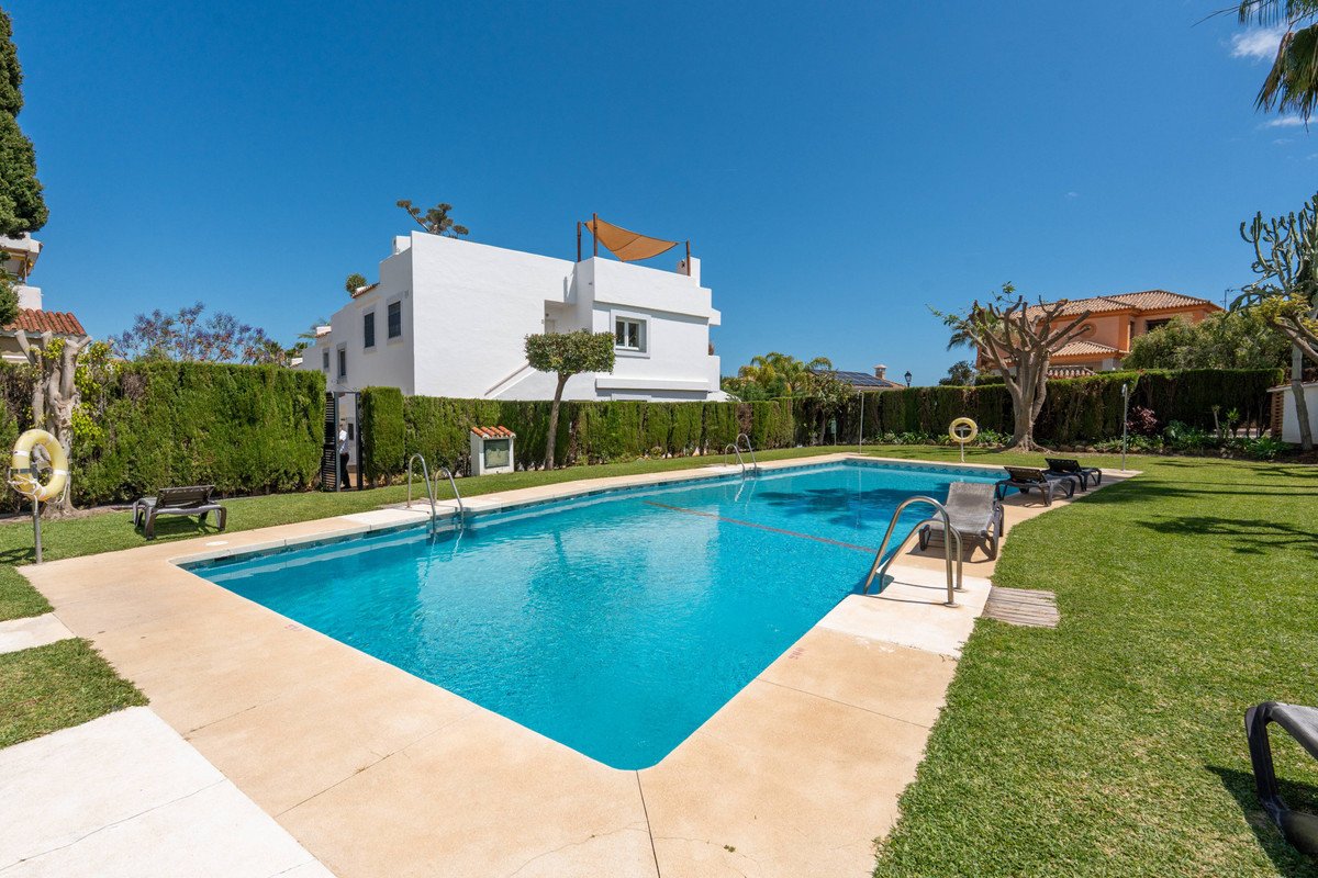 A bright blue swimming pool is surrounded by a manicured green lawn and bordered by a high green hedge. White residential buildings are visible in the background.
