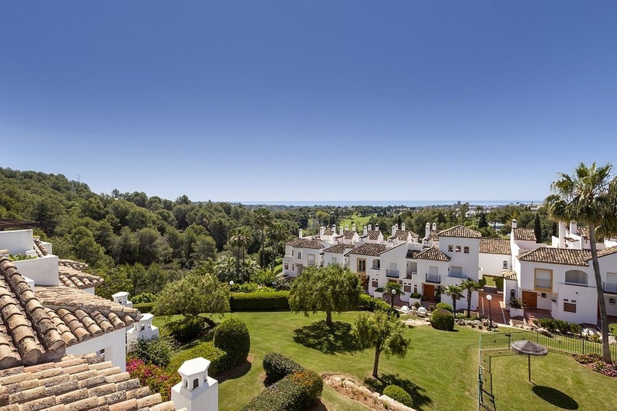 White village buildings with tiled roofs, green gardens, and sea view under blue sky