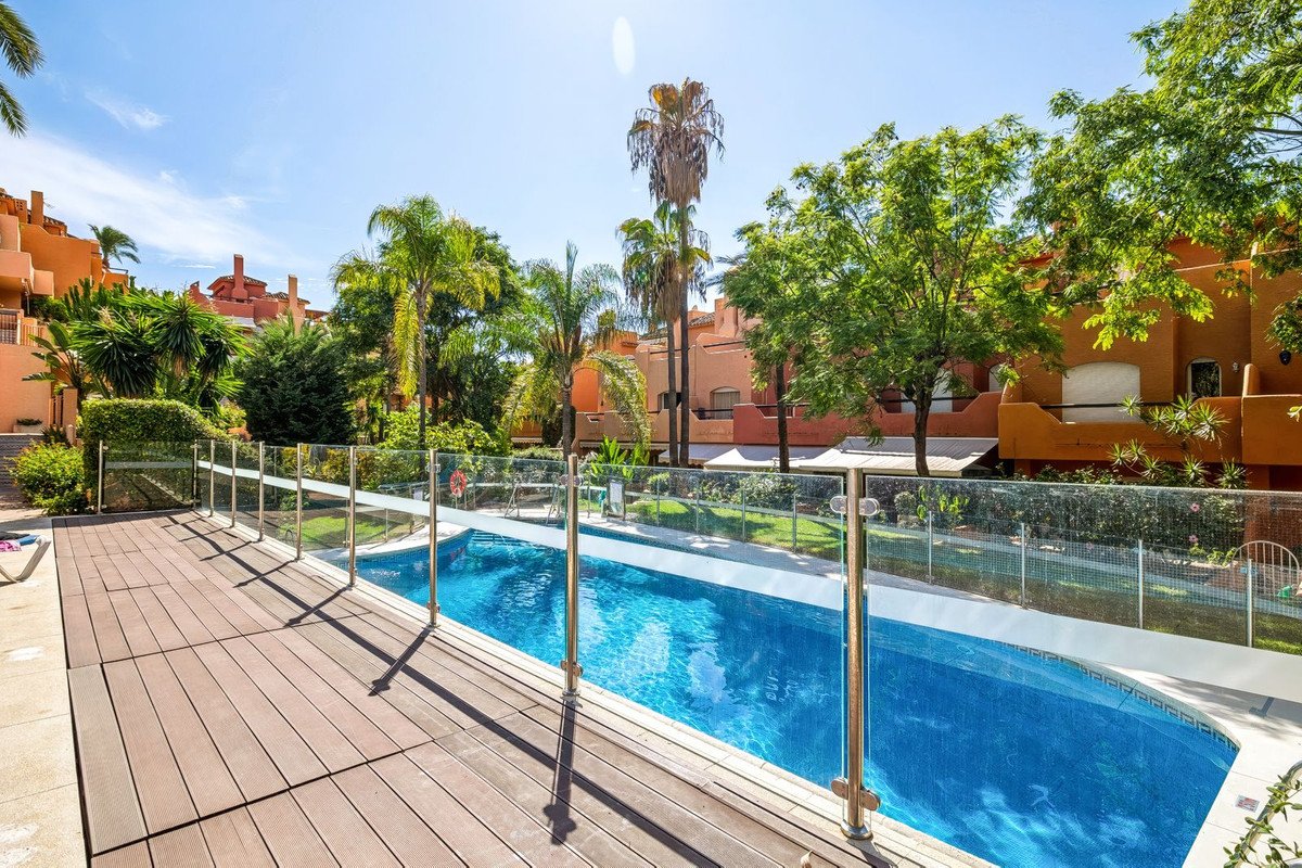 Outdoor swimming pool with a wooden deck and surrounding lush greenery, featuring palm trees and apartment buildings under a clear blue sky.