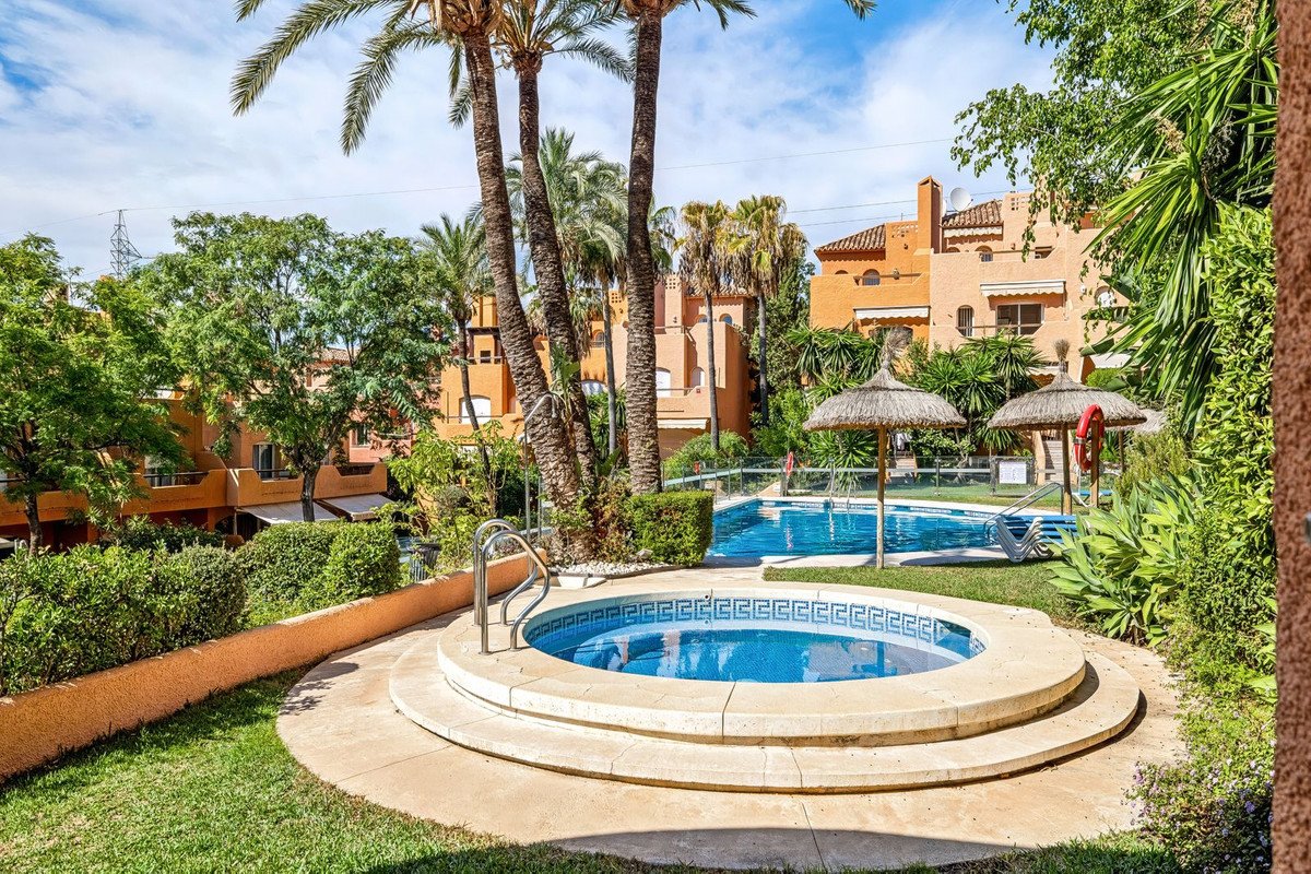 A round jacuzzi and a swimming pool surrounded by lush greenery and palm trees, with residential buildings in the background.