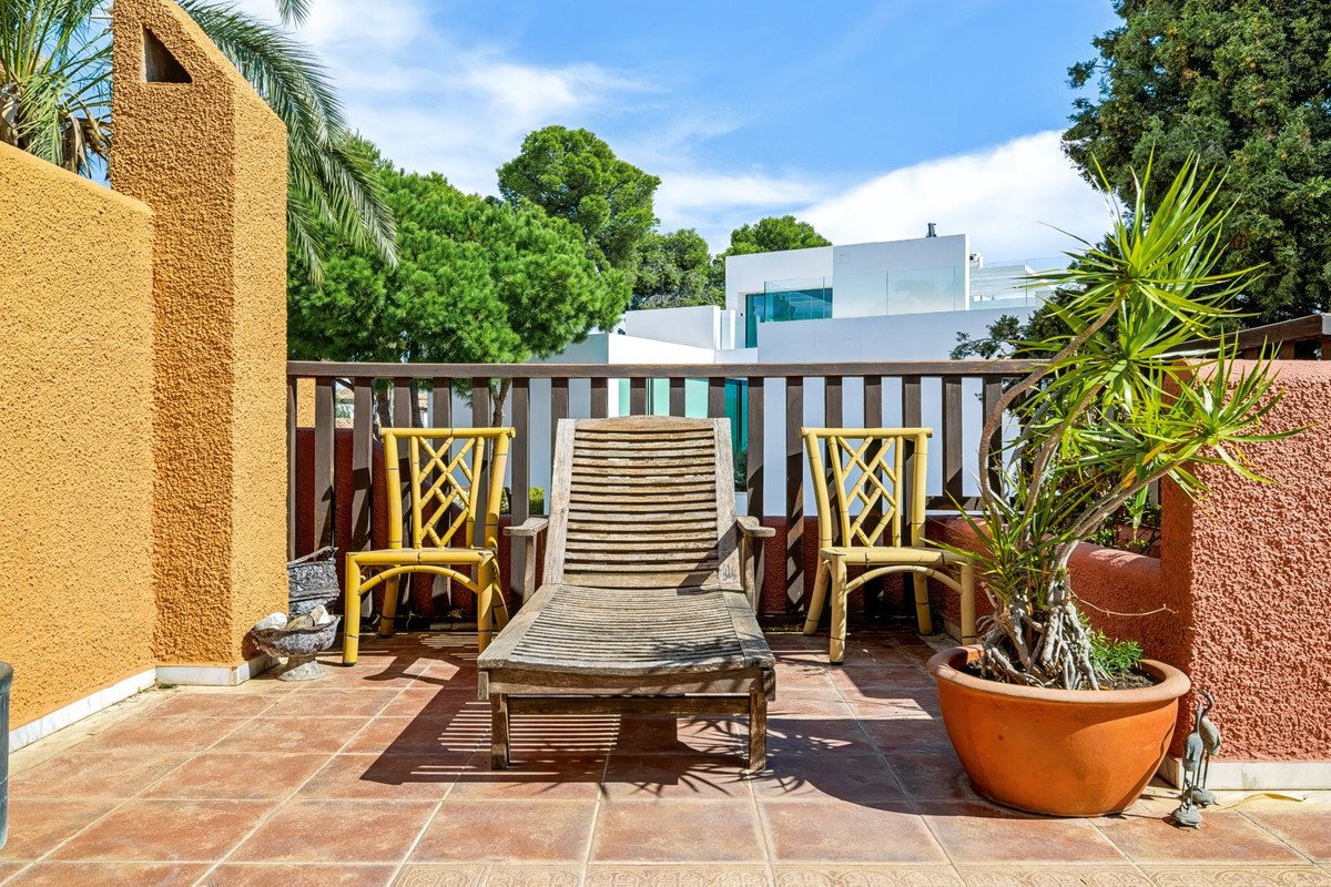 Terrace with wooden lounge chair, two yellow bamboo chairs, a potted plant, and a view of modern houses and trees.