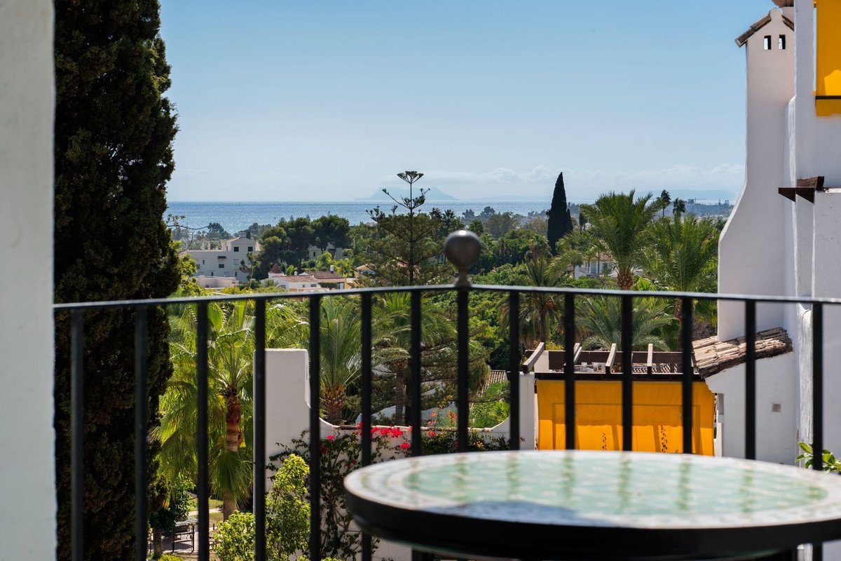 A balcony with a table offers a view of the sea, tropical trees, and white buildings, and a clear blue sky.