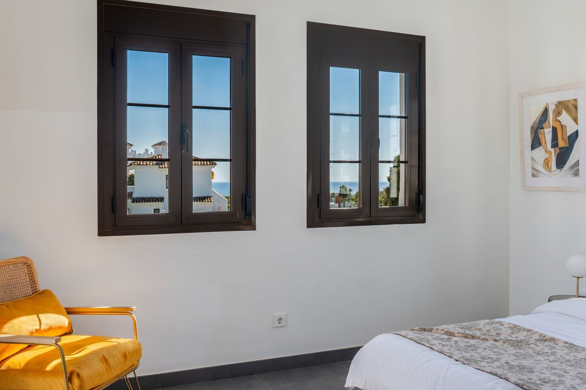 Bedroom with two windows offering a view of white buildings and the blue sea. Yellow armchair is visible.