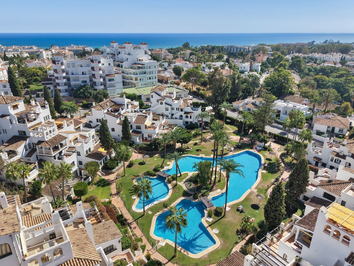 Aerial view of a residential complex with white buildings, swimming pools, lush gardens, palm trees, and the sea in the background.