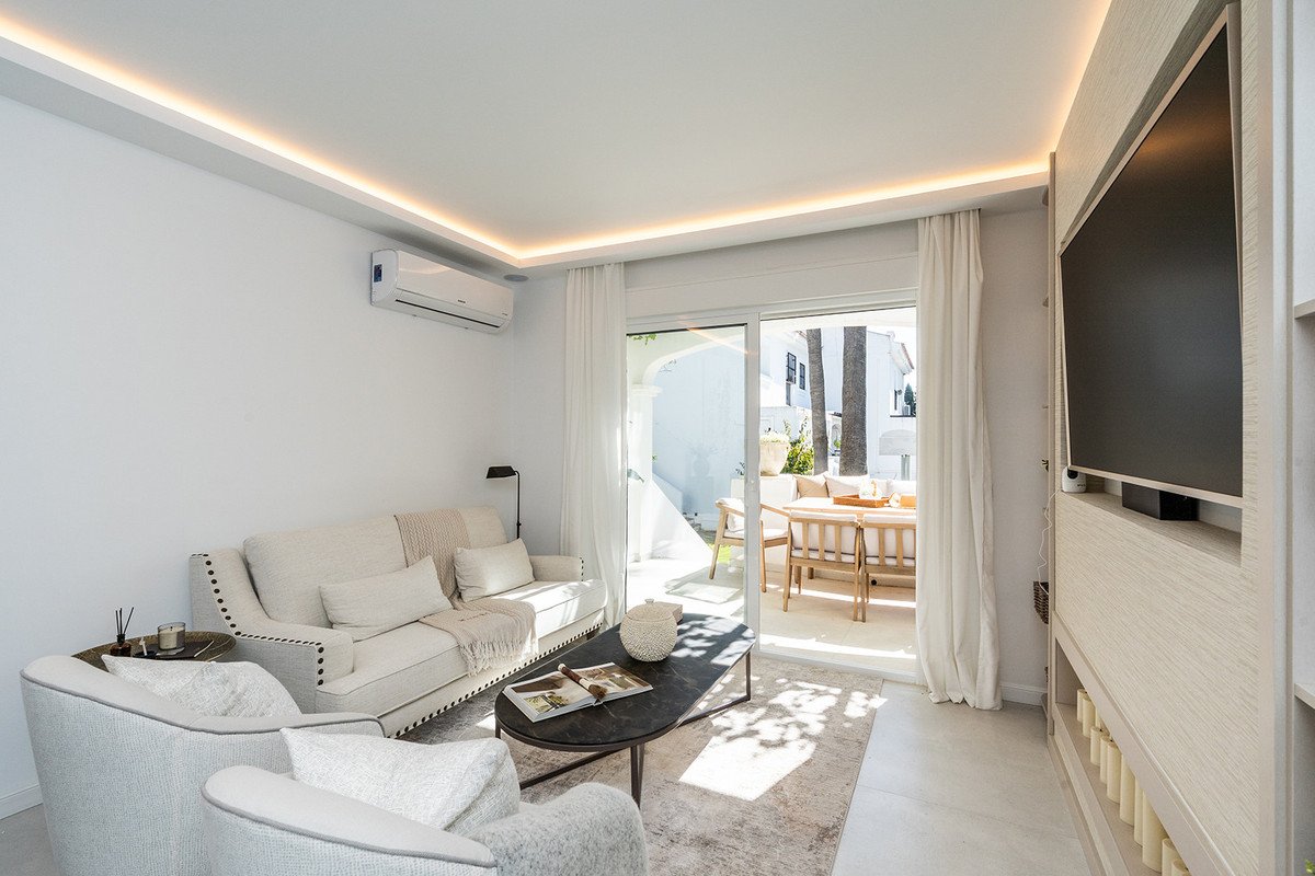 Modern living room with comfortable sofas, coffee table, TV, and a sliding door leading to a sunny terrace with outdoor dining.