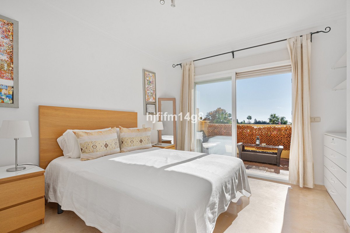 A bright bedroom with a large bed and sliding glass doors opening to a balcony with outdoor seating and a sunny view.