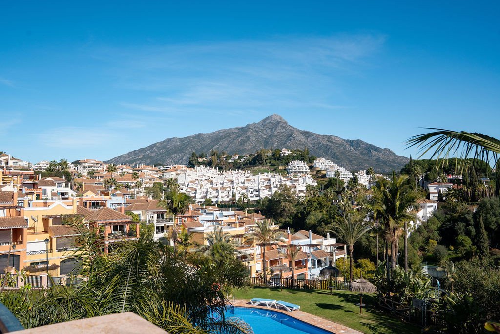 View of a village with white buildings and a prominent mountain in the background, featuring a swimming pool and palm trees.
