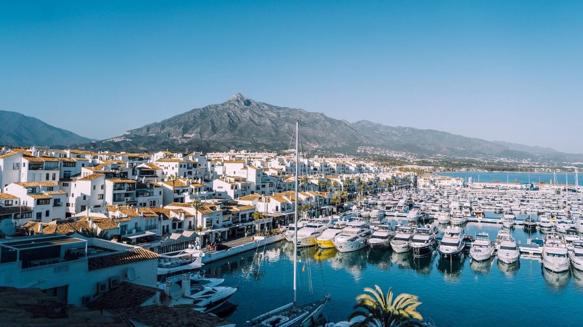 A scenic view of a bustling marina filled with yachts, overlooking a charming town and a majestic mountain range under a clear blue sky.