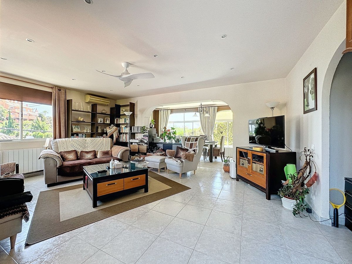 Spacious living and dining room area with large windows offering a green view, furnished with sofas, chairs, and a dining table.